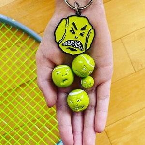 May include: A hand holding four yellow tennis balls with faces on them. The balls are all different expressions. There is a black metal keychain with a yellow tennis ball with a face on it that says "Ballgoyles.com".