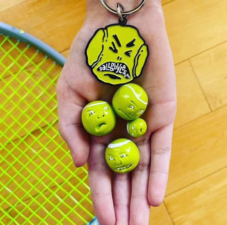 May include: A hand holding four yellow tennis balls with faces on them. The balls are all different expressions. There is a black metal keychain with a yellow tennis ball with a face on it that says "Ballgoyles.com".