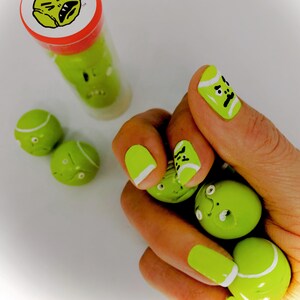 May include: A hand with green and white nail polish designed to look like tennis balls. The nails are decorated with black lines and a black dot to resemble a tennis ball's seam. There are also several green and white tennis balls in the hand.