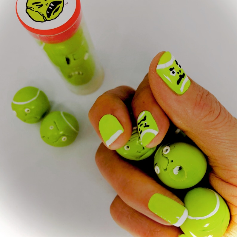 May include: A hand with green and white nail polish designed to look like tennis balls. The nails are decorated with black lines and a black dot to resemble a tennis ball's seam. There are also several green and white tennis balls in the hand.