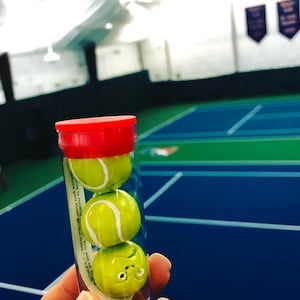May include: A clear plastic tube with a red lid contains three miniature tennis balls. The tube has a white label with the text "This is literally your "You win" of VENUS W..."