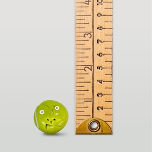 May include: A green and white tennis ball with a face sculpted on it. The ball is next to a wooden ruler with markings in inches.