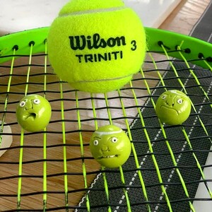 May include: A green tennis racket with three green tennis balls. One ball has the words "Wilson 3 TRINITI" printed on it. The other two balls have painted-on faces.