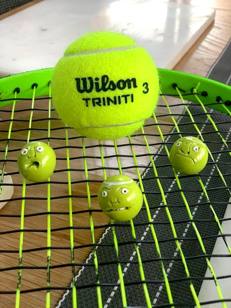 May include: A green tennis racket with three green tennis balls. One ball has the words "Wilson 3 TRINITI" printed on it. The other two balls have painted-on faces.