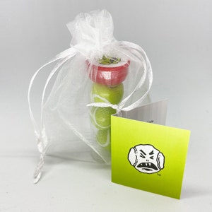 May include: A set of four small, round, green and red ornaments in a white organza bag. The ornaments have a cartoon face on them. The bag is tied with a white ribbon. A green card with a cartoon face and the word "TM" is in the foreground.