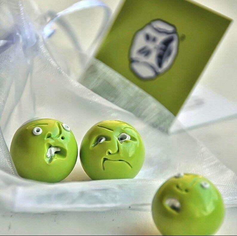 May include: Three green ceramic ornaments with different facial expressions. The ornaments are round and have a glossy finish. They are sitting on a white surface.