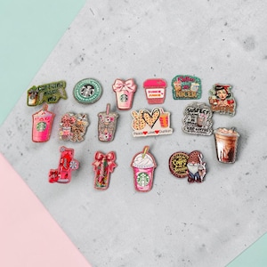 May include: A collection of small, decorative acrylic pins featuring various coffee-themed designs, including iced coffee cups, pink tumblers, a gnome, and text-based graphics like Dunkie Junkie and Peace Love Dunkin, all arranged on a grey surface.