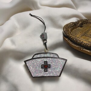 May include: White nurse hat charm with a red heart in the center of a black cross. The charm is made of glitter and has a black string attached to it.