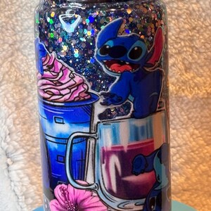 May include: A glitter-filled tumbler featuring a blue cartoon character, Stitch, with images of a drink and a flower. The tumbler has a black lid and sits on a light blue base. The design includes blue, pink, and purple colours.