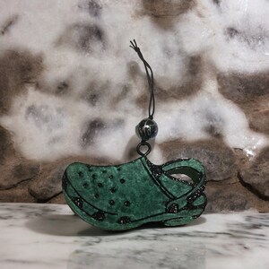 May include: A green and black glitter shoe charm in the shape of a clog. The charm has a black string with a black bead at the top.