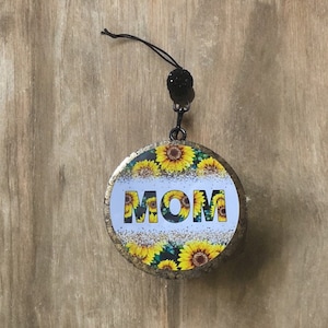 May include: Round, gold-colored ornament with a white background and a sunflower pattern. The word "MOM" is written in black letters on the ornament.
