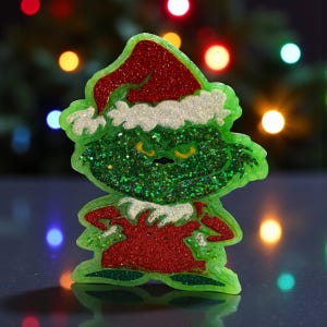 May include: A green glittery Christmas ornament in the shape of a cartoon character with a red Santa hat and red outfit. The ornament has a grumpy expression and is set against a blurred background of colorful lights.