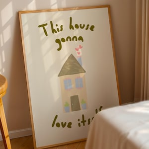 Könnte beinhalten: Ein gerahmter Kunstdruck mit einer skurrilen Illustration eines Hauses mit braunem Dach, blauen Fenstern und einem Schornstein mit Herzen. Der Text lautet "This house gonna love its". Das Kunstwerk befindet sich in einem hellen Rahmen.