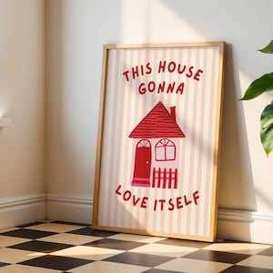 Könnte beinhalten: Ein gerahmter Kunstdruck mit einer roten Hausillustration und dem Text "THIS HOUSE GONNA LOVE ITSELF". Der Hintergrund hat rosa und weiße Streifen. Der Rahmen ist hellbraun. Der Druck lehnt an einer Wand.