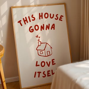 Könnte beinhalten: Ein gerahmter Kunstdruck mit cremefarbenem Hintergrund und rotem Text und Illustration. Der Text lautet "THIS HOUSE GONNA LOVE ITSEL" mit einer Zeichnung eines Hauses mit Herzen über dem Schornstein. Der Rahmen hat eine helle Holzfarbe.
