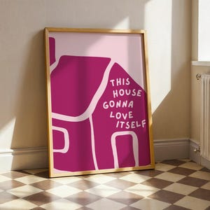 Könnte beinhalten: Ein gerahmter Kunstdruck mit einer magenta Hausillustration und dem weißen Text „THIS HOUSE GONNA LOVE ITSELF“. Der Hintergrund ist hellrosa. Der Holzrahmen umgibt das Kunstwerk. Der Druck lehnt an einer Wand.