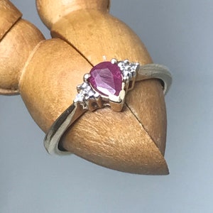 以下が含まれることがあります： 中央に洋梨型の濃い赤色の宝石、両側に小さな透明な石が配置されたシルバーリング。リングのセッティングはゴールドのようです。リングは木製の関節式の手の上に展示されています。