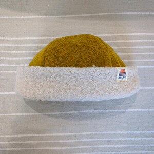 Può includere: Un berretto giallo senape con fodera in sherpa bianca e un piccolo patch con logo che dice "DOLPO" sul lato.