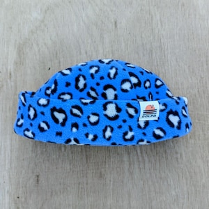 Peut inclure: Un bonnet en polaire bleu avec un motif léopard blanc et noir. Le chapeau a un bord replié et une petite étiquette blanche avec le mot "DOLPO" imprimé dessus. Le chapeau est fait d'une matière douce et chaude.