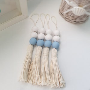 May include: Four white and light blue wooden bead tassels with cream coloured yarn. The tassels are hanging from a white surface.