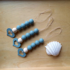 May include: Three teal blue wooden bead garlands with white wooden heart charms and twine hanging loops. The garlands are displayed on a light brown wooden surface with a white seashell.