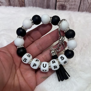 Op de afbeelding: Een ronde sleutelhangerarmband met afwisselend matzwarte en witte ronde kralen, gescheiden door kleine zilverkleurige strass-tussenstukjes. Vijf witte kubuskralen met zwarte letters vormen het woord SPURS, vergezeld van een zwarte kwast.