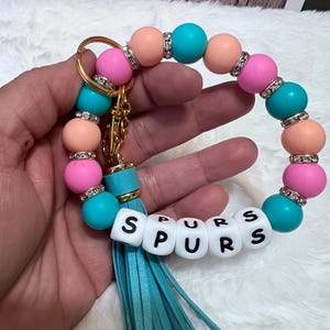 Op de afbeelding: Een ronde sleutelhangerarmband met afwisselende kralen in perzik, felroze en turquoise. Elke kraal wordt gescheiden door een kleine zilverkleurige strass-tussenring. Witte vierkante kralen vormen het woord SPURS. Een turquoise kwastje hangt aan een goudkleurige metalen ring.