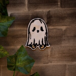 May include: A white ghost with black eyes and a small mouth, embroidered on a fabric patch. The ghost has two small feet and is floating in the air.
