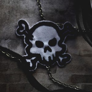 May include: A white and black embroidered skull and crossbones patch attached to a silver-toned metal chain and a black fabric ribbon. The patch features a stylized skull with dark eye sockets and a nose cavity, outlined in thick black thread.
