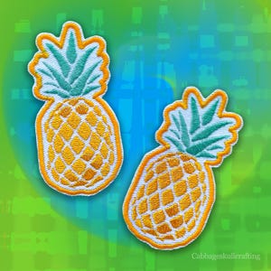 Puede incluir: Dos parches bordados de piña con cuerpos de fruta amarillos con patrón de cuadrícula, hojas verdes y un borde naranja grueso. Los parches son idénticos en diseño, mostrando costuras detalladas y colores vibrantes sobre una base blanca.