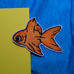 May include: A close-up of an orange embroidered fish patch on a blue fabric background. The fish has a large eye and a detailed fin.