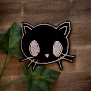 May include: Black embroidered iron-on patch of a cartoon cat with white eyes and whiskers.