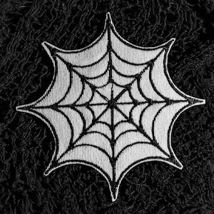 May include: A monochrome embroidered spiderweb patch. The patch has a detailed web design with a central point and radiating lines, set against a textured black background. Perfect for adding a spooky touch to clothing or accessories.