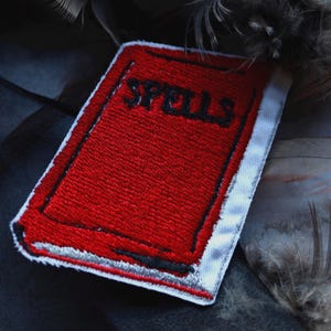 May include: A rectangular embroidered patch featuring a bright red book cover with the word SPELLS stitched in black thread. The patch has a white border and black outlines defining the book shape, with a silver-coloured spine detail at the bottom.