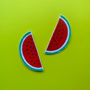 May include: Two red watermelon slices with green rinds and black seeds on a bright yellow background.