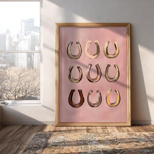Puede incluir: Una l&aacute;mina de arte enmarcada que presenta una cuadr&iacute;cula de nueve ilustraciones de herraduras en varios tonos de marr&oacute;n, beige y dorado, sobre un fondo rosa empolvado s&oacute;lido. El marco rectangular es de madera clara con un perfil sencillo.