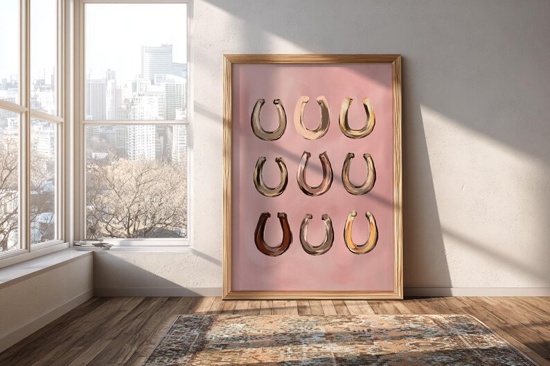 Puede incluir: Una l&aacute;mina de arte enmarcada que presenta una cuadr&iacute;cula de nueve ilustraciones de herraduras en varios tonos de marr&oacute;n, beige y dorado, sobre un fondo rosa empolvado s&oacute;lido. El marco rectangular es de madera clara con un perfil sencillo.