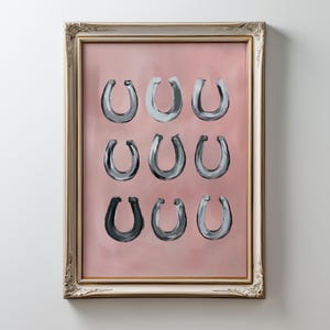 Puede incluir: Una l&aacute;mina de arte enmarcada que presenta nueve ilustraciones de herraduras pintadas dispuestas en una cuadr&iacute;cula de tres por tres. Las herraduras est&aacute;n representadas en varios tonos de gris y negro sobre un fondo rosa empolvado s&oacute;lido.