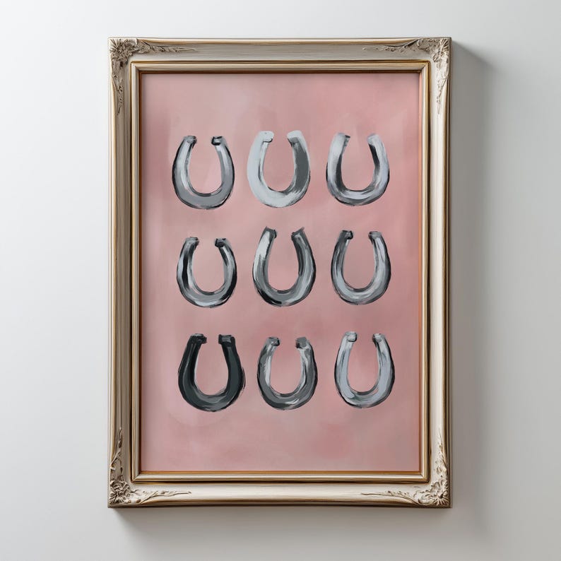 Puede incluir: Una l&aacute;mina de arte enmarcada que presenta nueve ilustraciones de herraduras pintadas dispuestas en una cuadr&iacute;cula de tres por tres. Las herraduras est&aacute;n representadas en varios tonos de gris y negro sobre un fondo rosa empolvado s&oacute;lido.