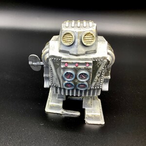 May include: A silver wind-up robot toy with a round head, two large round eyes, and four small round gauges on its chest. The robot has a key on the side and a small arm that moves.