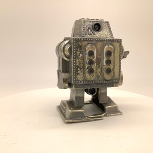 May include: A silver-colored robot toy with a worn, distressed finish. The robot has a round head with a single eye and a small, rectangular body with multiple round details. The robot is standing on a white surface.