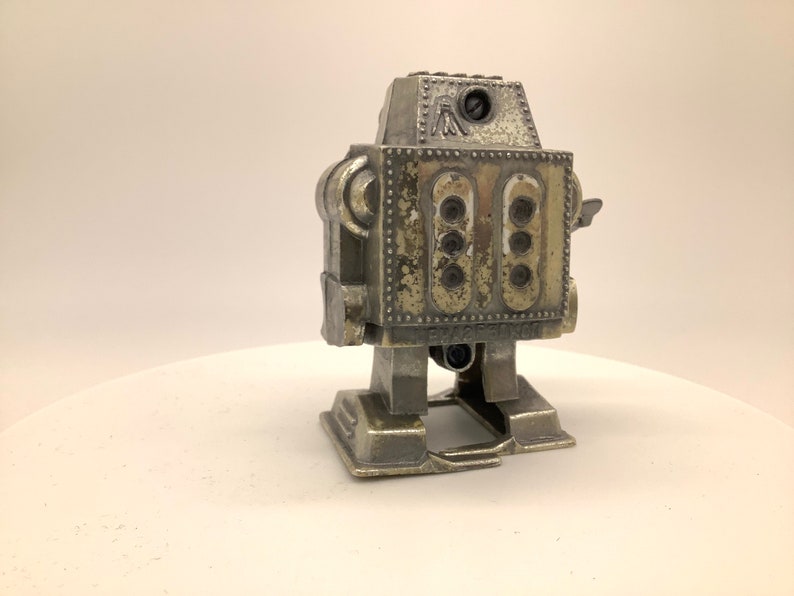 May include: A silver-colored robot toy with a worn, distressed finish. The robot has a round head with a single eye and a small, rectangular body with multiple round details. The robot is standing on a white surface.