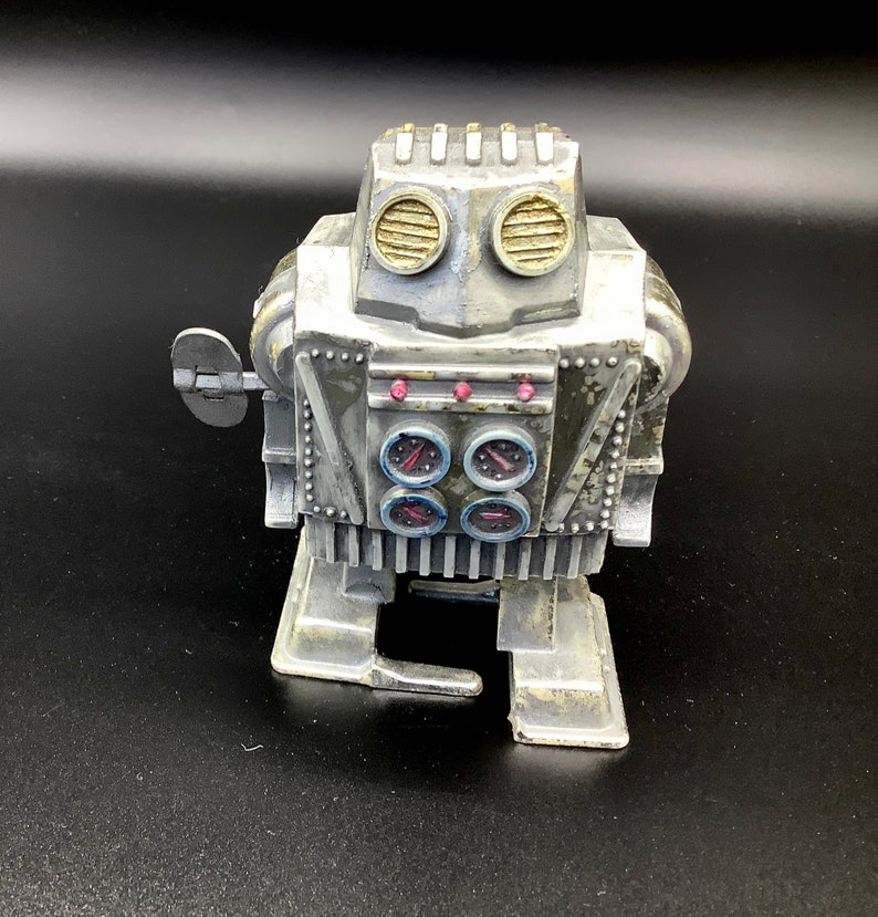 May include: A silver wind-up robot toy with a round head, two large round eyes, and four small round gauges on its chest. The robot has a key on the side and a small arm that moves.