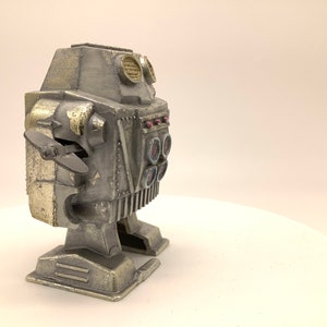 May include: A silver and gold robot toy with a propeller on the side and a row of round buttons on the front. The robot has a weathered, vintage look.