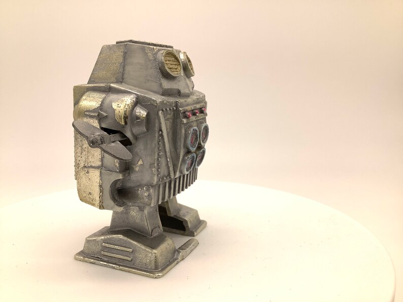 May include: A silver and gold robot toy with a propeller on the side and a row of round buttons on the front. The robot has a weathered, vintage look.
