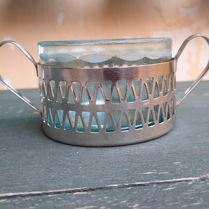 May include: Silver sugar bowl with a glass insert and two handles. The bowl has a decorative pattern of triangles around the rim.