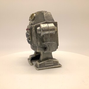 May include: A silver robot toy with a rounded head and a rectangular body. The robot has two small, round eyes and a small, round antenna on top of its head. The robot has two arms and two legs, and it is standing on a white surface.