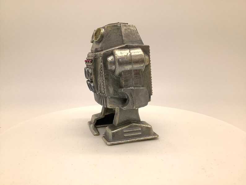 May include: A silver robot toy with a rounded head and a rectangular body. The robot has two small, round eyes and a small, round antenna on top of its head. The robot has two arms and two legs, and it is standing on a white surface.