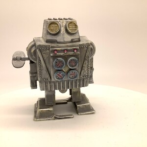May include: A silver wind-up robot toy with a round head, four round gauges, and a single arm with a round disc. The robot has a silver body with black and red details.