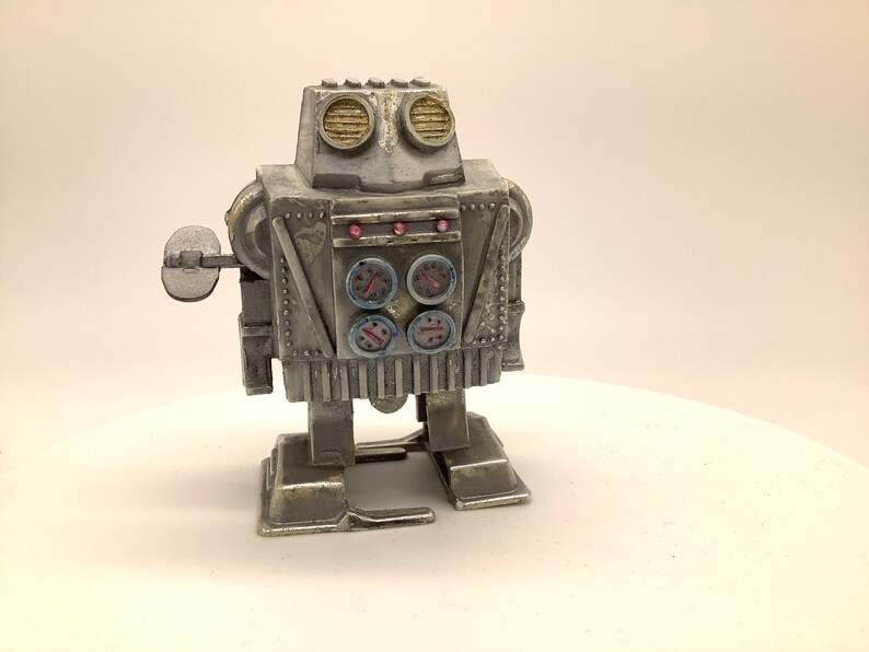 May include: A silver wind-up robot toy with a round head, four round gauges, and a single arm with a round disc. The robot has a silver body with black and red details.